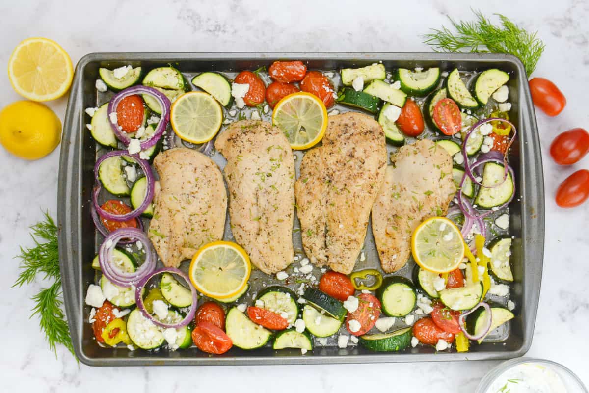 The finished Greek sheet pan meal. The chicken is surrounded by vegetables and topped with feta. To the side of the sheet pan sit a bowl of tzatziki, cherry tomatoes, dill and a cut lemon