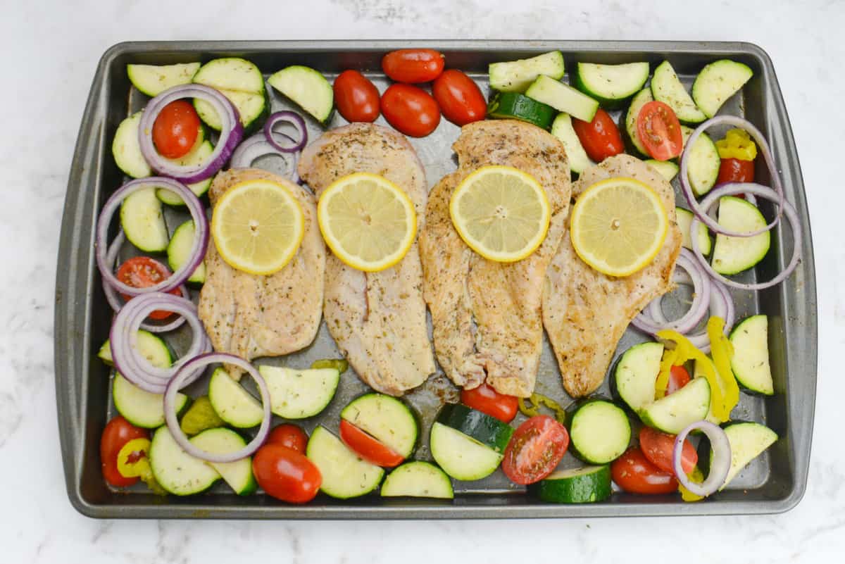 The assembled sheet pan meal ready for baking. The vegetables surround the seared chicken which is topped each with one lemon slice 