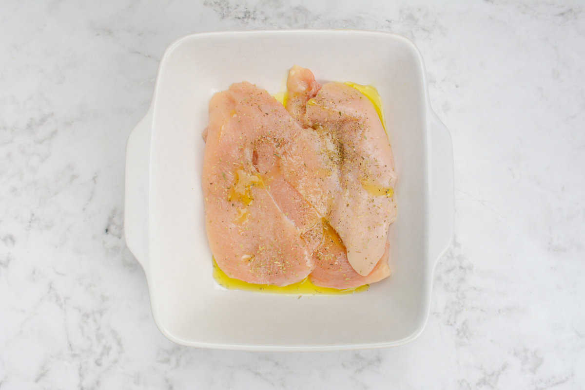 A square dish with raw chicken covered in olive oil, salt, oregano and pepper