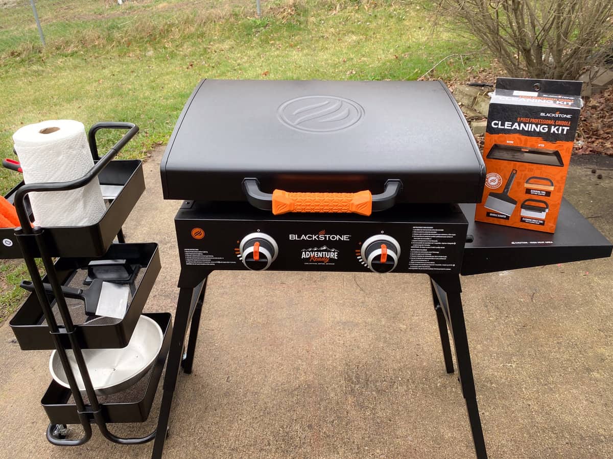 A blackstone grill sits on the patio. On the left side is a small accessory cart. To the right is a Blackstone cleaning kit box.