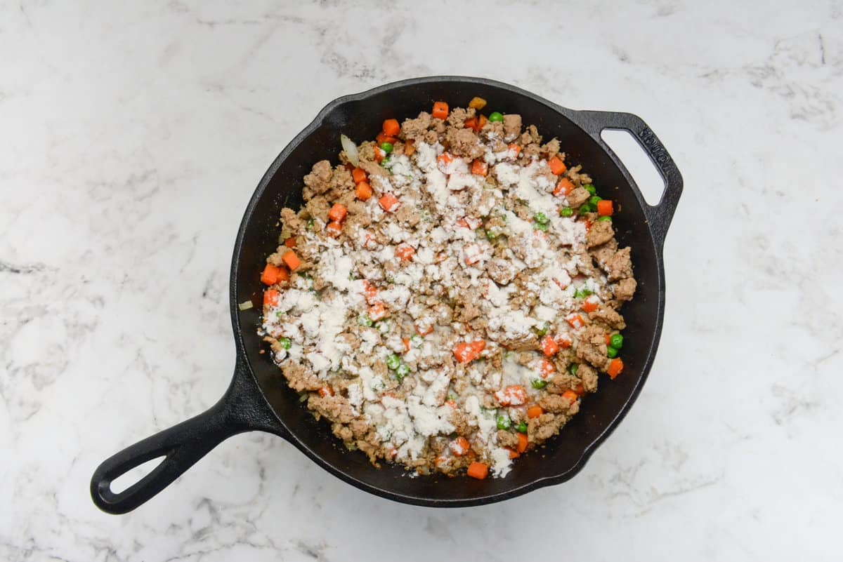 A cast iron skillet with the meat and vegetable mixture. Flour is sprinkled on top but not mixed in.