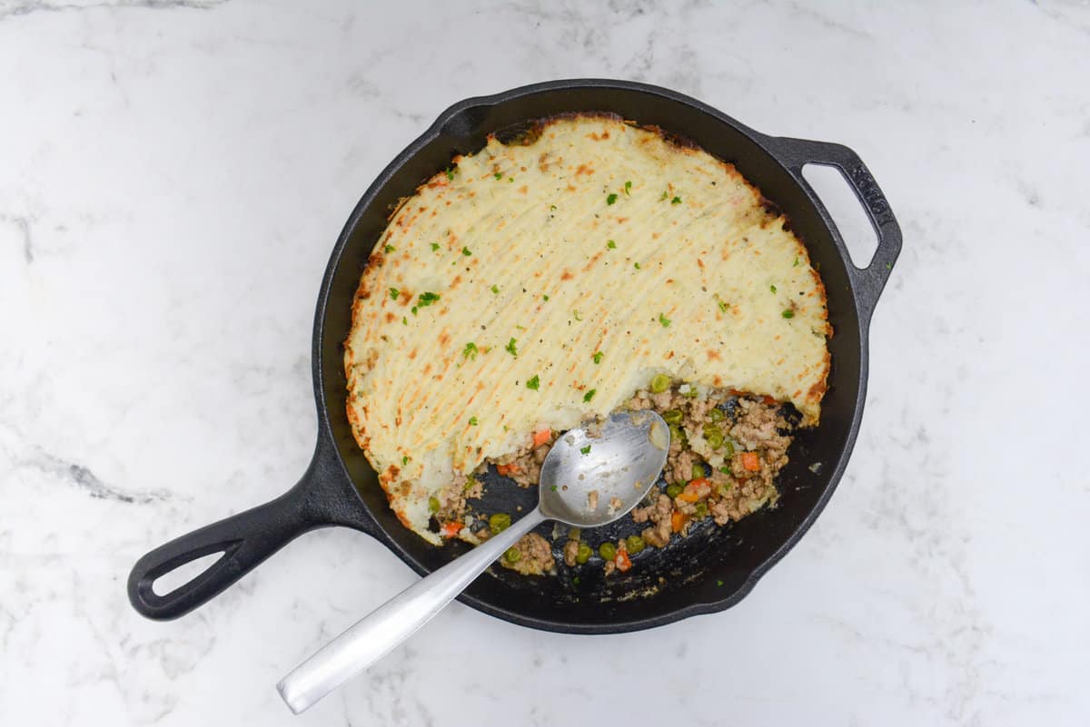 The cooked Turkey Shepherd's Pie with a scoop taken out. A silver serving spoon sits inside the skillet where small amounts of vegetables and meat peek out from under the potatoes.