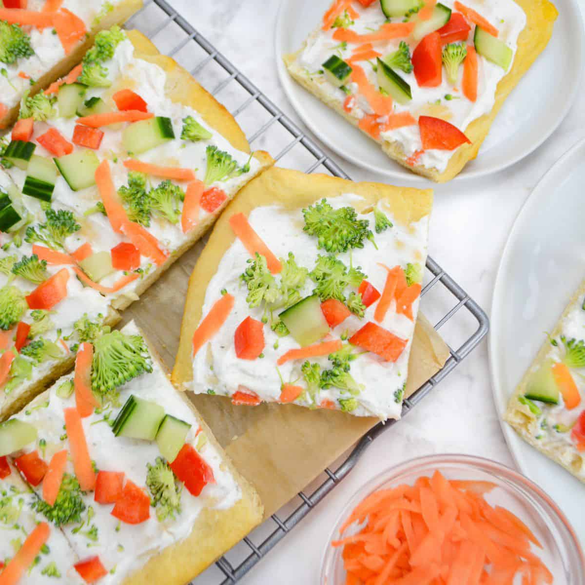 Veggie pizza cut into slices sits on top of a cooling rack lined with parchment. Two small plates with slices surround it and a small bowl of shredded carrots.