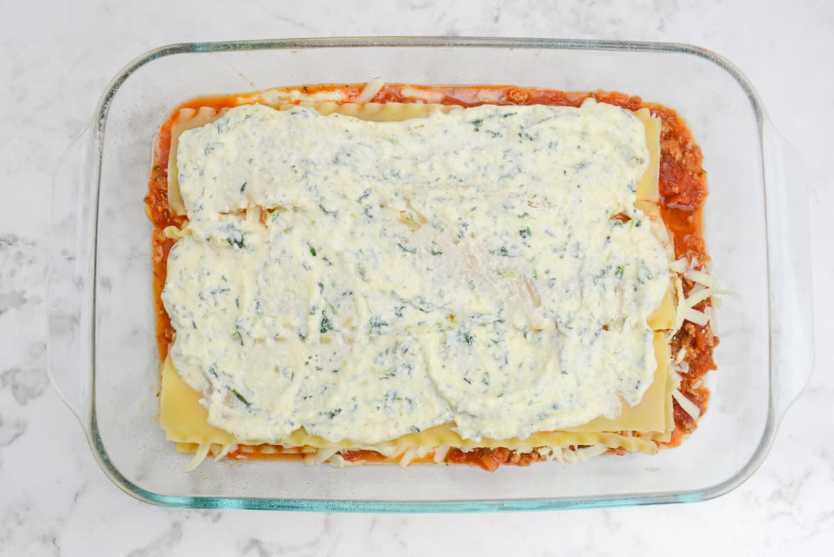 A clear baking dish is filled with various layers of lasagna. The current top layer has a white and green cottage cheese layer