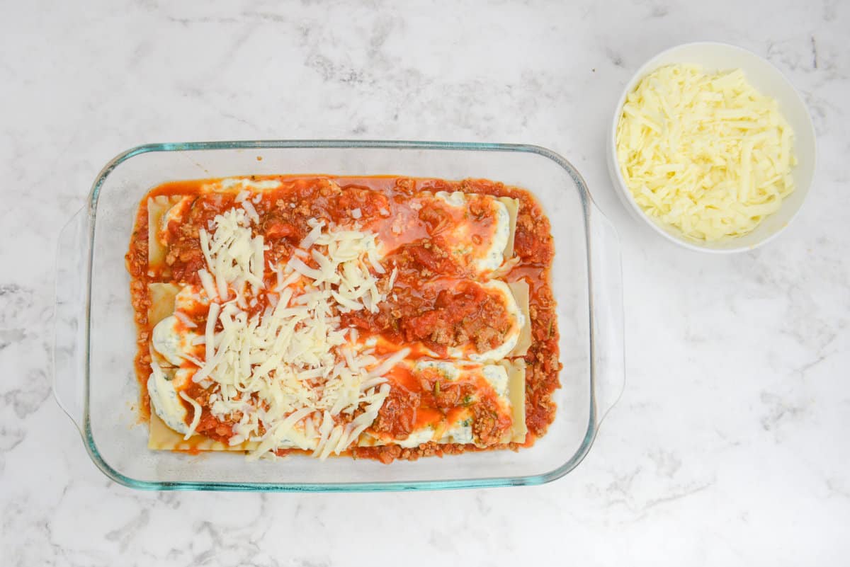 A clear baking dish is filled with the assembled lasagna and cheese is partially covering the noodles. A small bowl of mozzarella sits to the side.