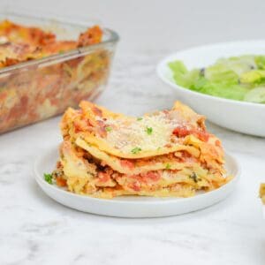 Single serving of baked lasagna on a small white plate. A bowl of salad sits to the right and the casserole dish to the left.
