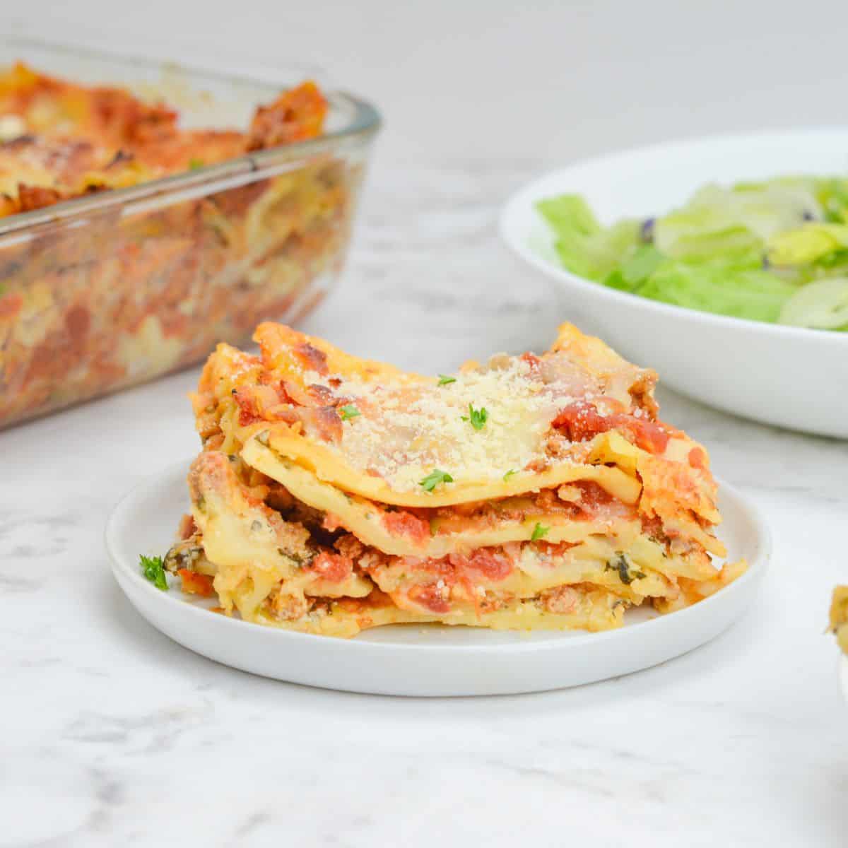 A single serving of baked lasagna sits on a white plate. A bowl of salad sits to the right and the casserole dish to the left.