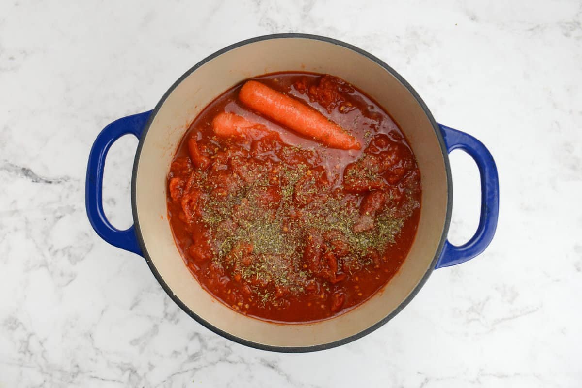 A blue dutch oven is filled with tomato sauce. Dried herbs sit on top prior to mixing along with two carrots