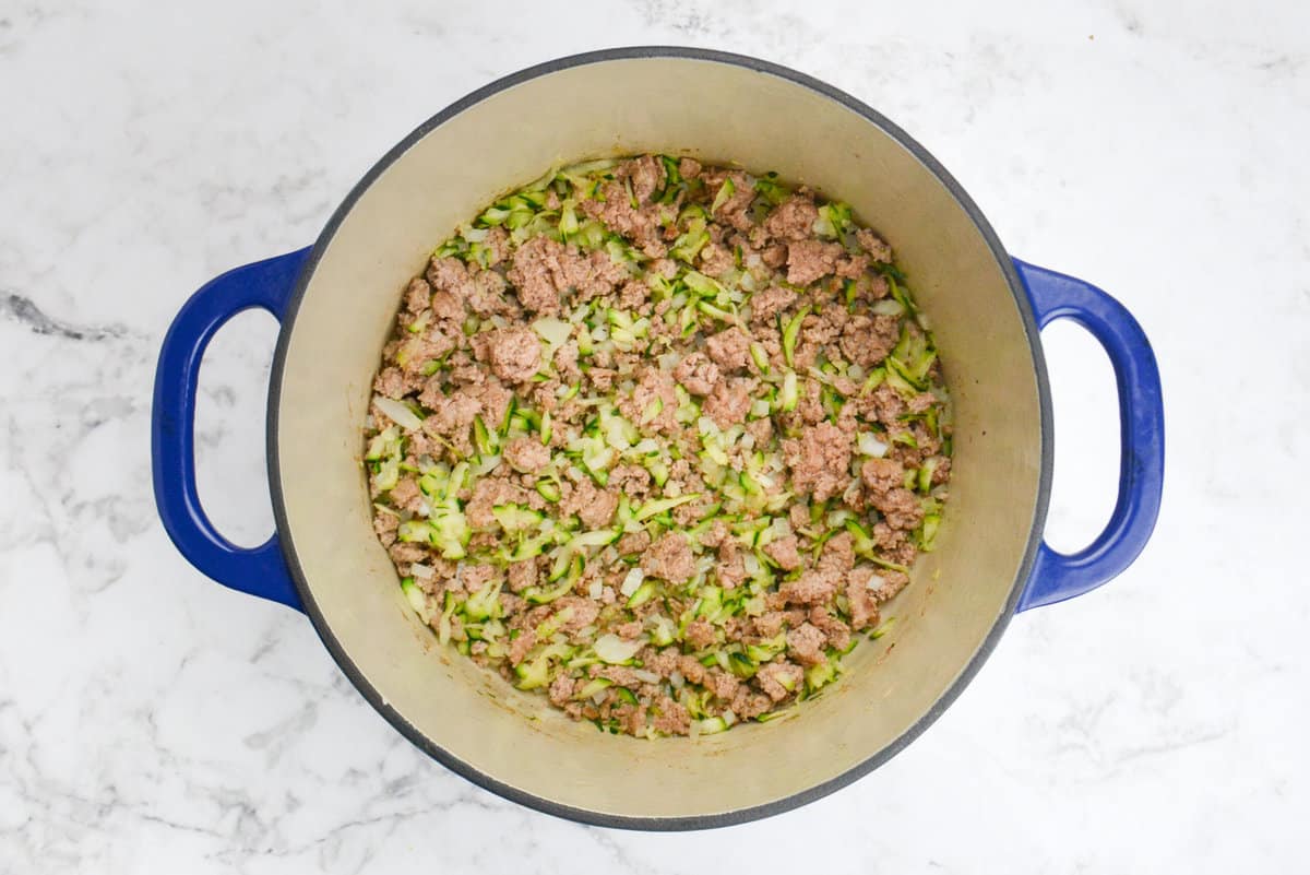 A blue dutch oven is filled with browned turkey and dotted with shredded zucchini