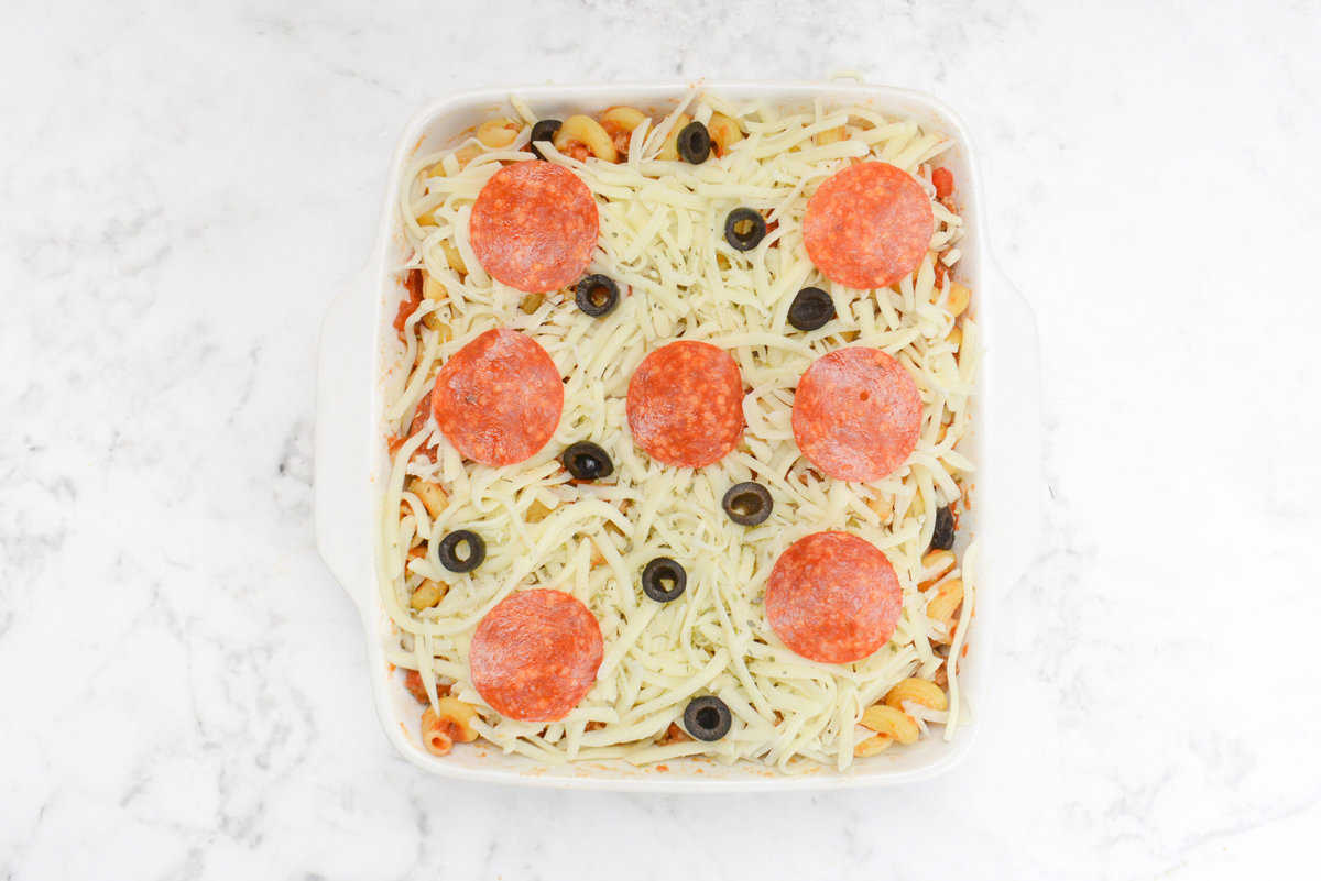 A white square casserole dish topped with cheese and whole slices of pepperoni and scattered sliced olives 