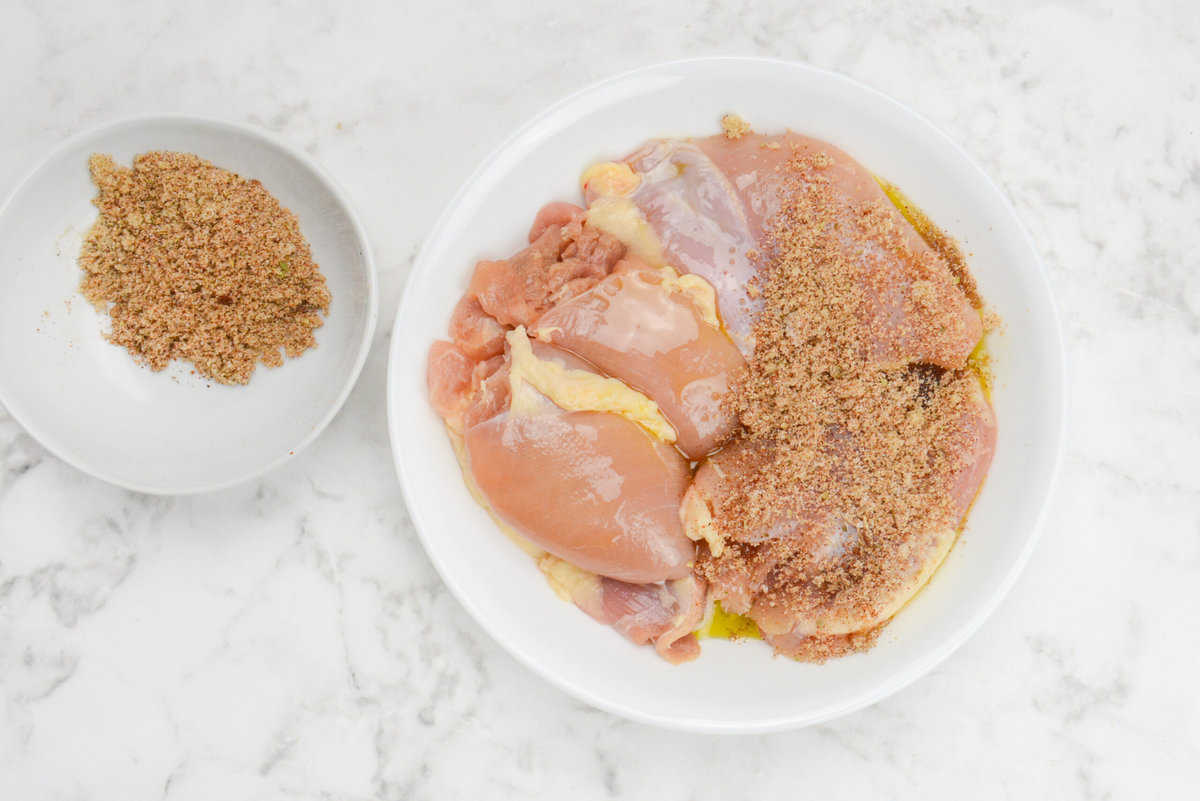 A plate of raw chicken thighs with seasoning sprinkled over half of the chicken. The remaining seasoning sits in a bowl to the side 