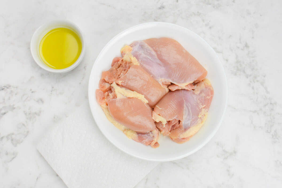 A plate of raw chicken thighs. To the side sides a small bowl of olive oil and a folded paper towel