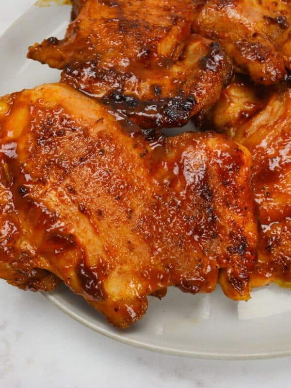 Close up of a white plate with two bbq chicken thighs. Sauce coats the chicken and the edges are browned and caramelized