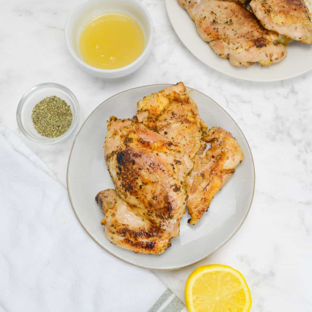 A small plate with two cooked chicken thighs. A small bowl of oregano, a bowl of honey, a white napkin and a sliced lemon sit to the side. A plate of chicken thighs sits to the far right. 