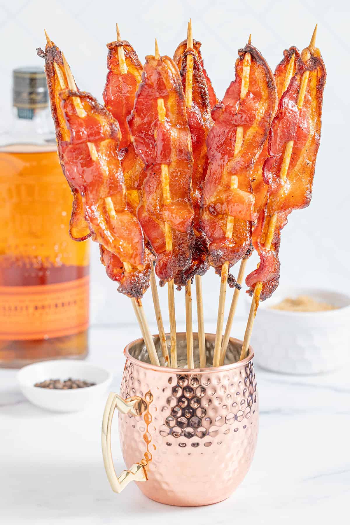 Candied bacon strips on skewers in a metal cup on a table.