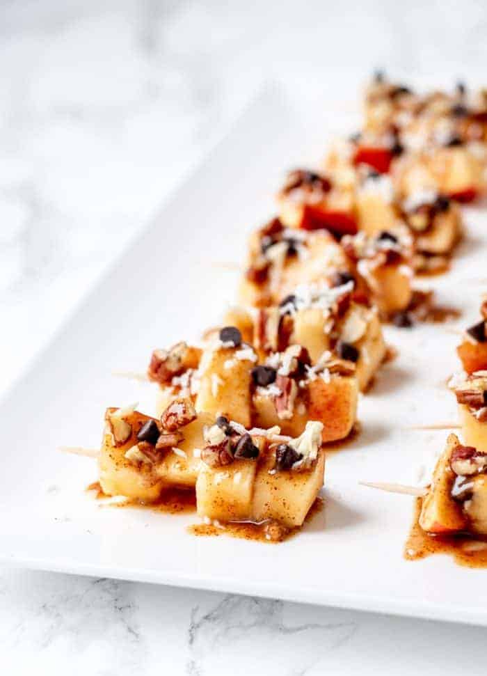 Apple pieces, chocolate chips, and nuts on toothpicks on a rectangular plate.
