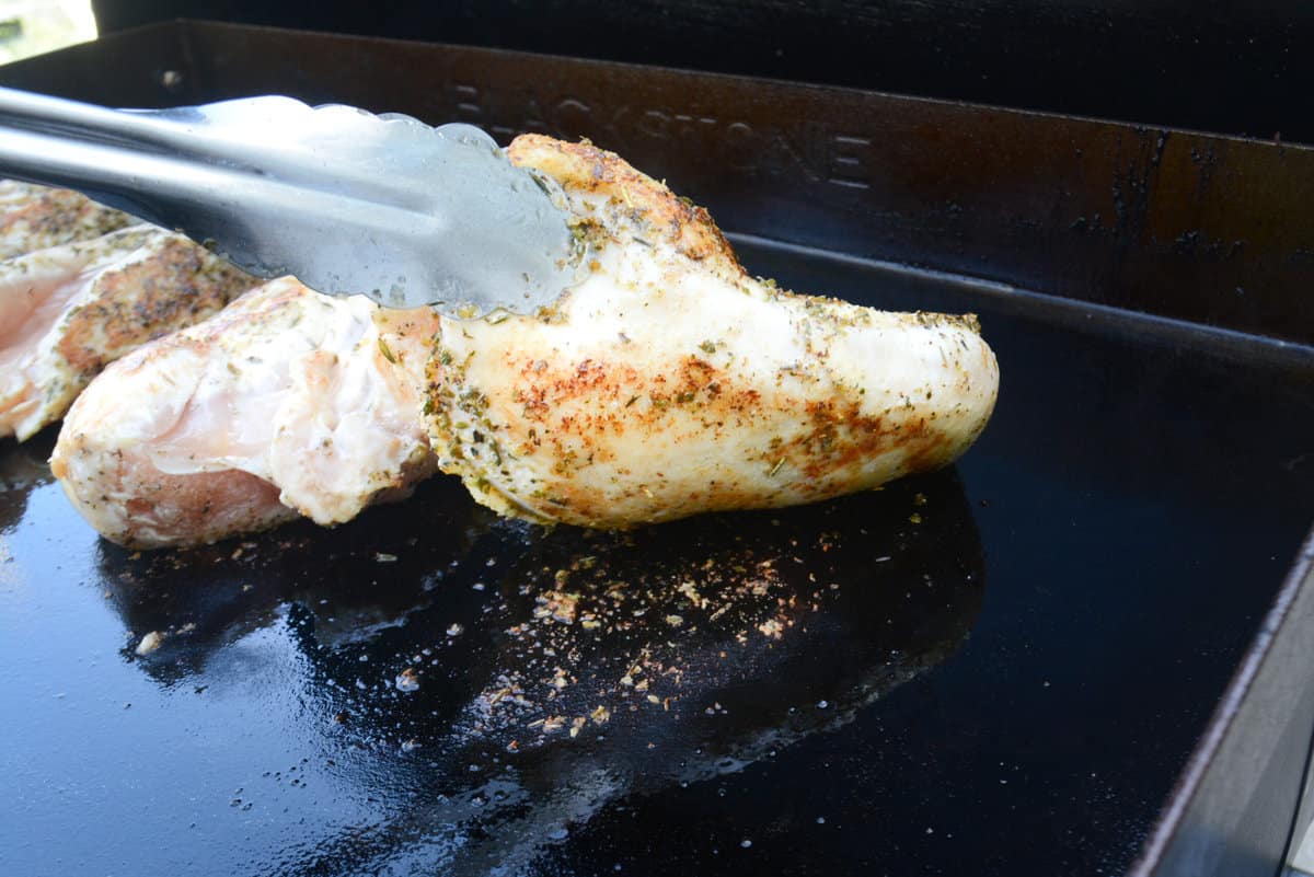 Silver metal tongs lift one edge of a single piece of chicken breast showing how it lifts easily