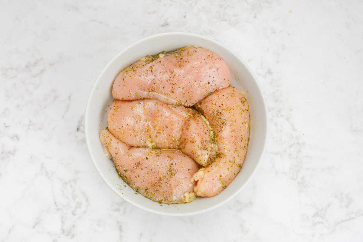 A large white bowl containing raw chicken that is covered in olive oil and seasoning
