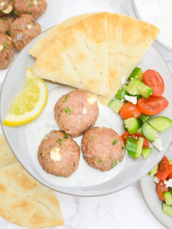 An overhead view of a plate filled with baked meatballs sitting in a spoonful of tzatziki sauce. To the side is a lemon slice, pita, and a cucumber and tomato salad. In the background is an additional plate with the same ingredients, more pita and leftover meatballs.