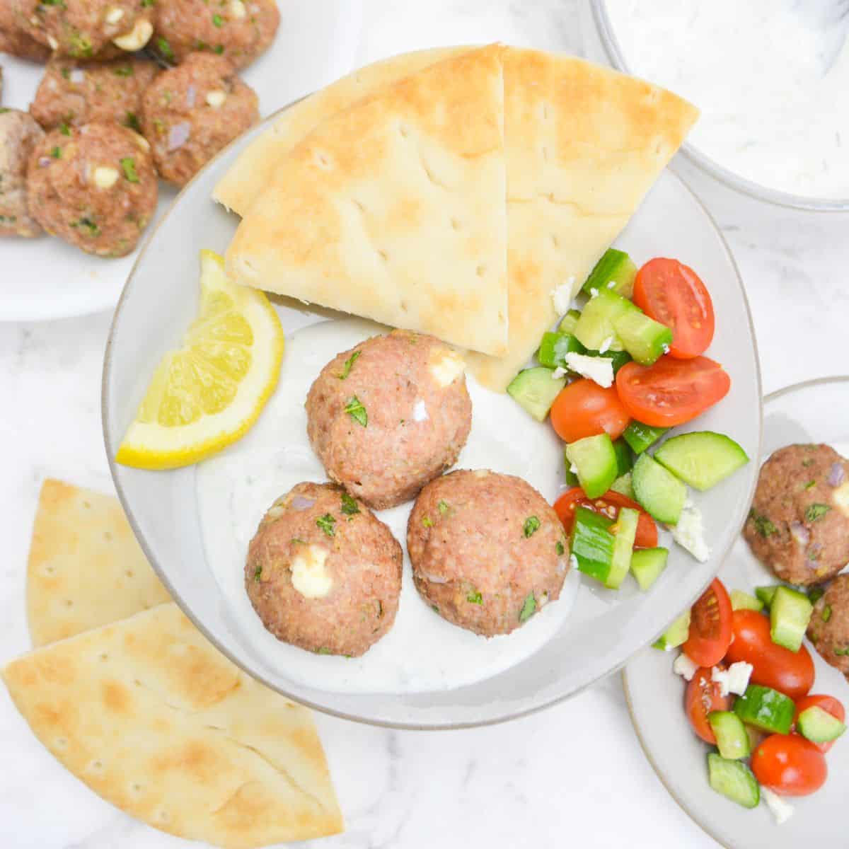 An overhead view of a plate filled with baked meatballs sitting in a spoonful of tzatziki sauce. To the side is a lemon slice, pita, and a cucumber and tomato salad. In the background is an additional plate with the same ingredients, more pita and leftover meatballs.