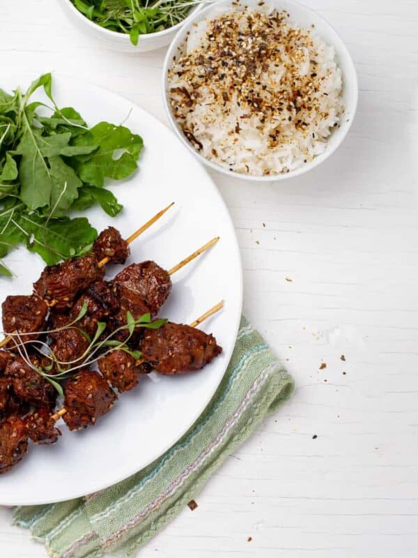 A plate of Green Tea Beef Kebabs with leafy greens beside a plate of seasoned rice.