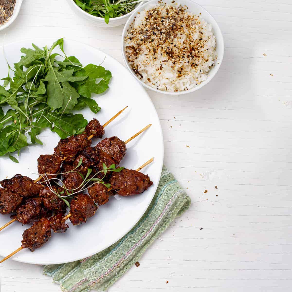 A plate of Green Tea Beef Kebabs with leafy greens beside a plate of seasoned rice.