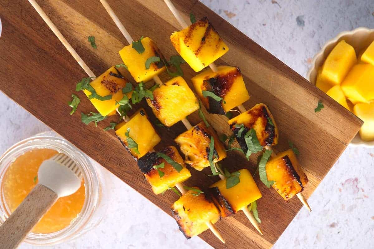Grilled mango cubes on skewers on a wooden board on a counter.