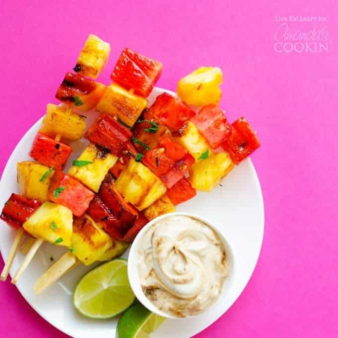 Pineapple and watermelon pieces on wooden skewers on a plate with a dipping sauce and lime slices.