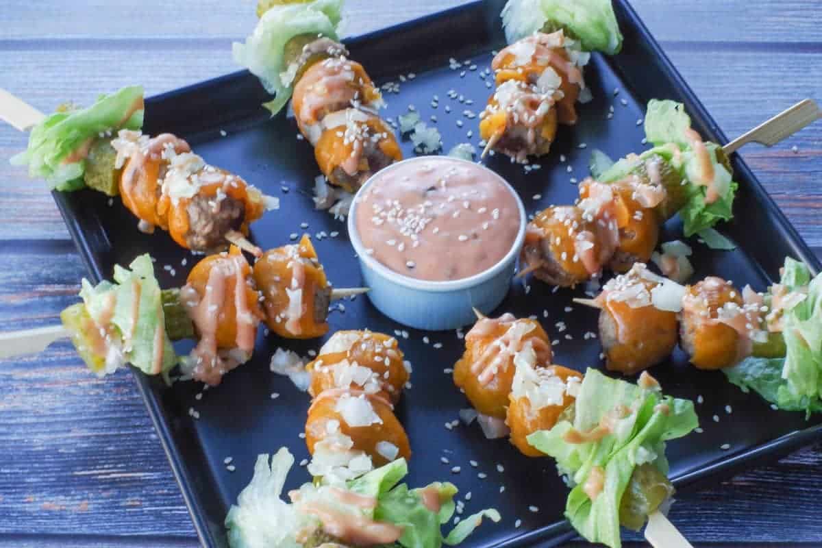 Cheese-covered meatballs, pickles, onions, and lettuce on skewers on a plate with a dipping sauce.