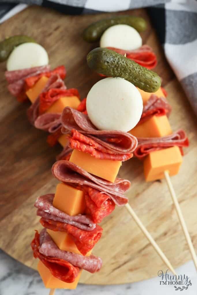 Pepperoni slices, mozzarella balls, cheese, and pickles on a skewers on a wooden board.