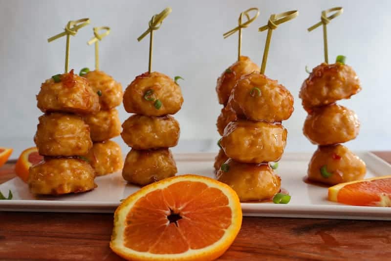 Coated chicken balls on wooden sticks on a serving tray with orange slices on it.