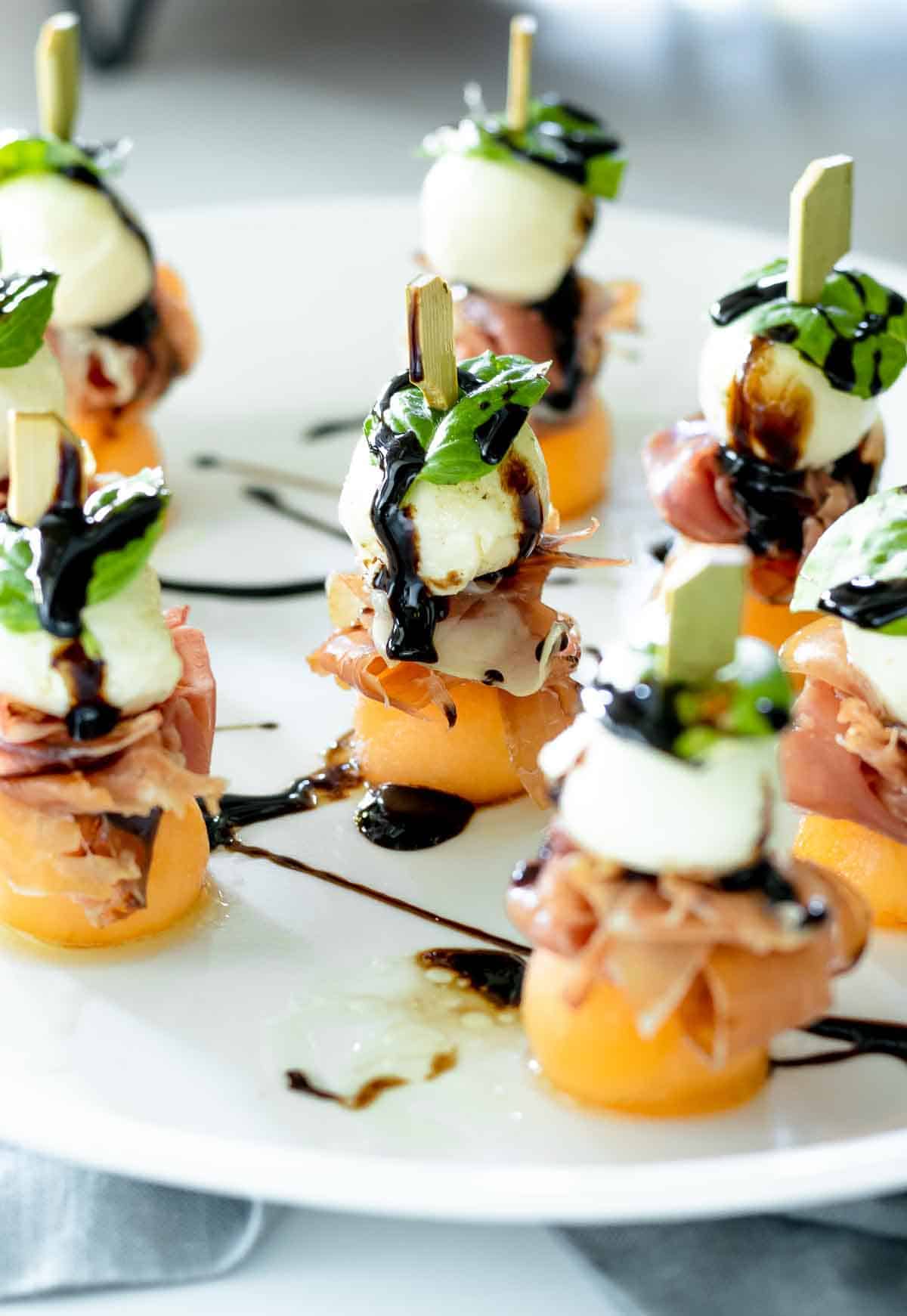 Porsciutto, melon, and mozzarella balls on skewers, covered in balsamic vinegar on a plate.