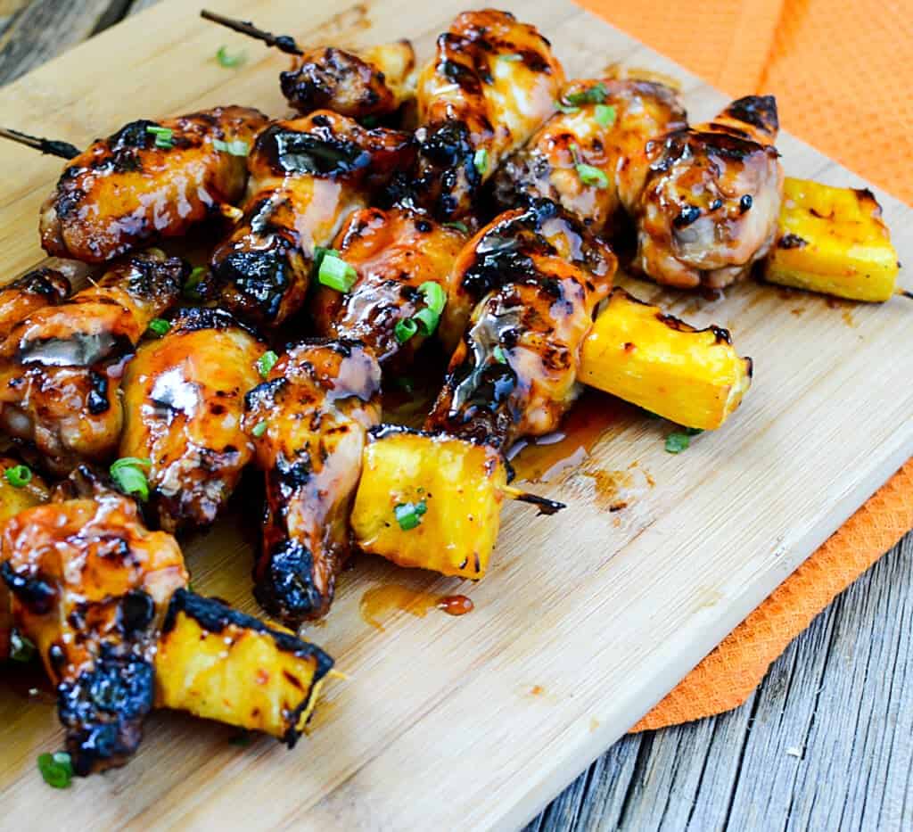Chicken wings and pineapples with sauce and hers on skewers on a wooden board on a table.