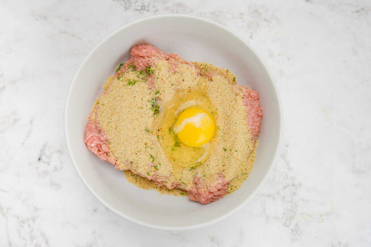 A large white bowl filled with raw turkey. Breadcrumbs are sprinkled on top and an egg is cracked into the center but not yet mixed.