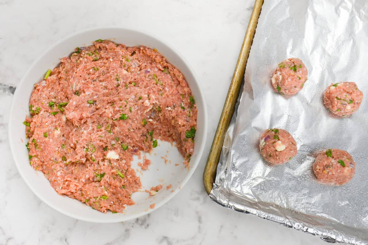 A large white bowl is filled with the Greek turkey meatball mixture and scoops taken out. To the side is a foil lined baking sheet with four formed meatballs.