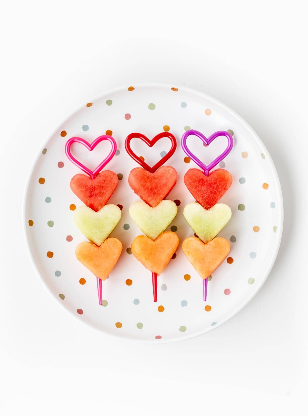 Heart-shaped watermelon, honeydew, and cantaloupe melons on heart-shaped skewers on a dotted plate.