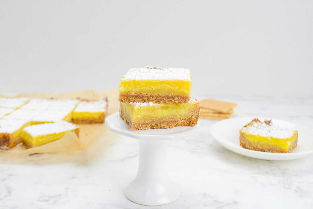 A small single cupcake stand with two sliced lemon bars placed on top. In the background are a plate with one lemon bar, a stack of graham crackers and a sheet of parchment with the remaining sliced bars.