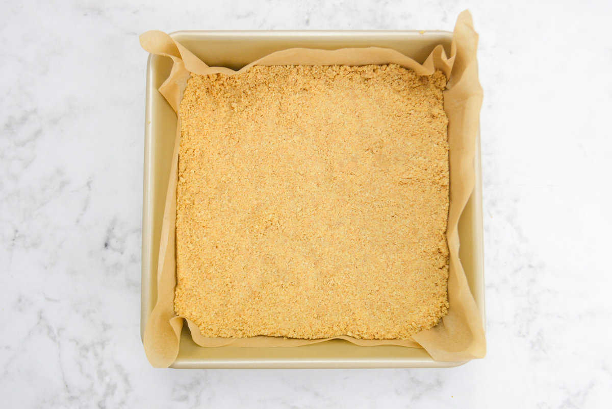 A gold square baking pan. The pressed crust sits in the pan while the brown parchment hangs slightly over the sides
