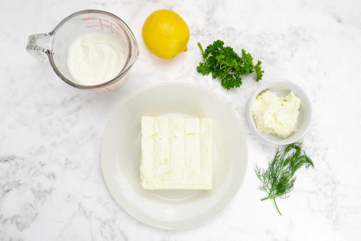 The ingredients needed to make the dip are shown. A plate with a full bock of feta, a measuring cup with yogurt, a small bowl with cream cheese sit alongside herbs and a whole lemon.