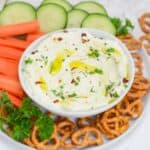 A bowl of whipped feta dips sits on a serving plate. Surrounding the bowl is pretzels, and fresh sliced vegetables for dipping