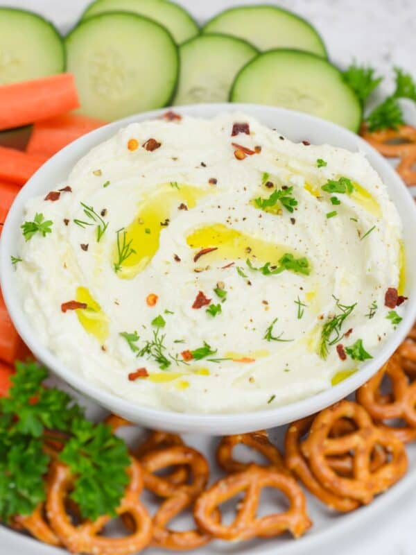 A bowl of whipped feta dips sits on a serving plate. Surrounding the bowl is pretzels, and fresh sliced vegetables for dipping