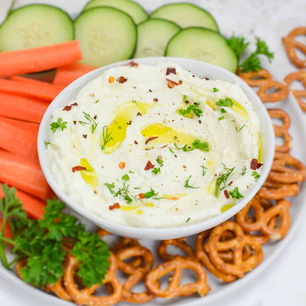 A close up of a white bowl filled with the finished whipped feta dip. On top of the dip are flakes of crushed red peppers, a light drizzle of oil and chopped herbs. The bowl sits on top of a plate filled with fresh cut veggies and pretzels. 