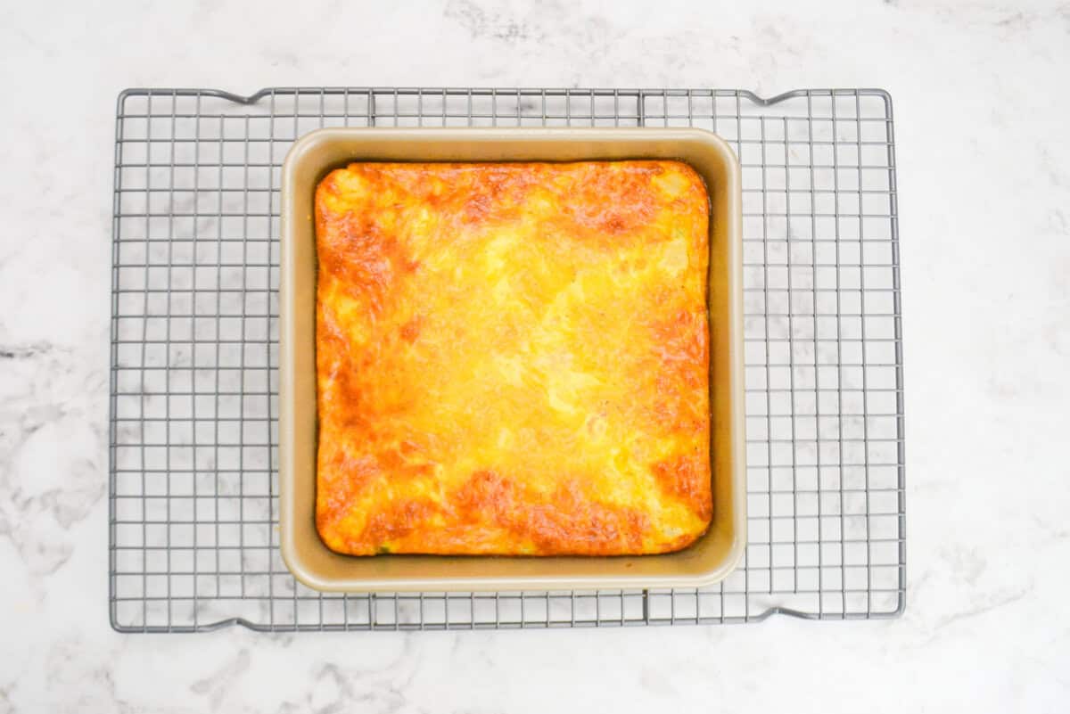 The baked breakfast casserole in the baking pan sits on top of a silver cooling rack. the edges are browned with the center slightly less browned 