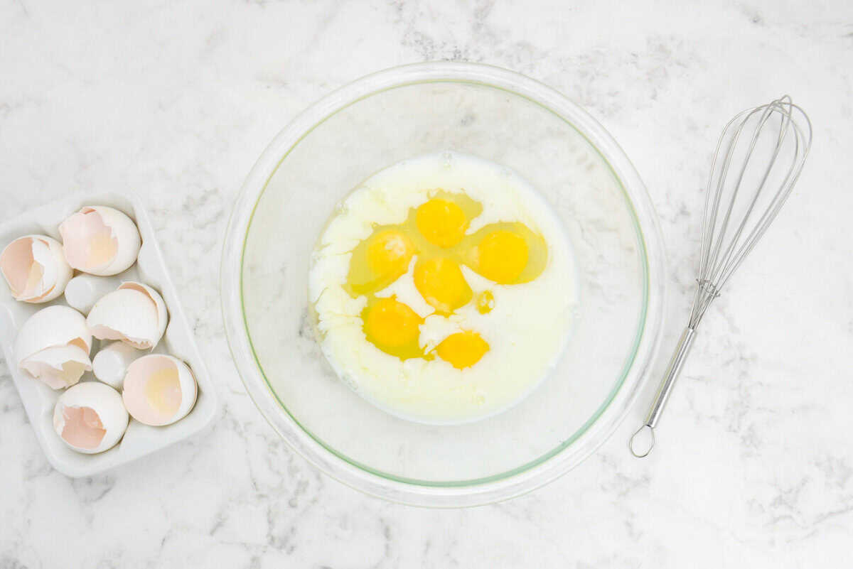 A large clear mixing bowl filled with unbeaten eggs and milk. To the side is a whisk and the cracked egg shells 
