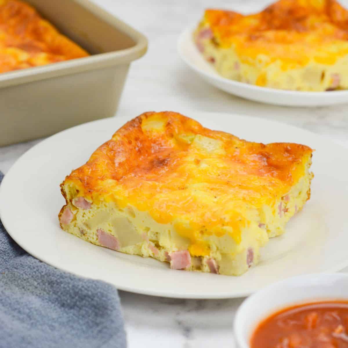 A slice of baked weight watchers breakfast casserole sits on a white plate. To the side is a small plate with a single slice, the baking pan, a gray napkin and a small bowl of salsa 