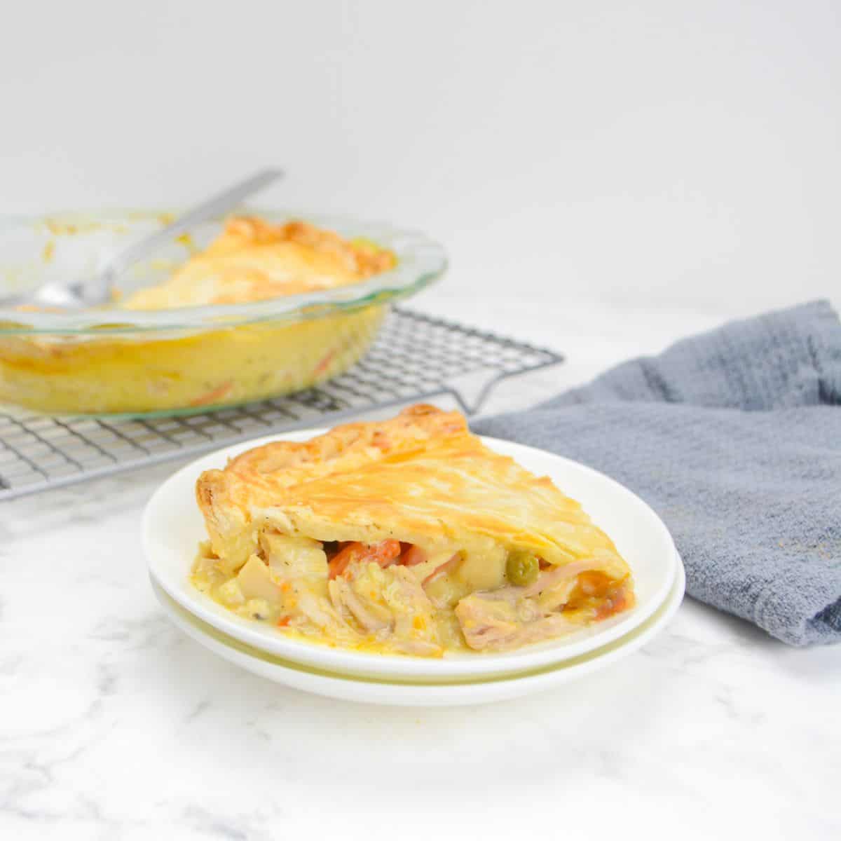 Single slice of Weight Watchers Chicken Pot pie sits on top of a white plate next to a grey napkin. In the background is a cooling rack with the casserole dish and a large spoon.