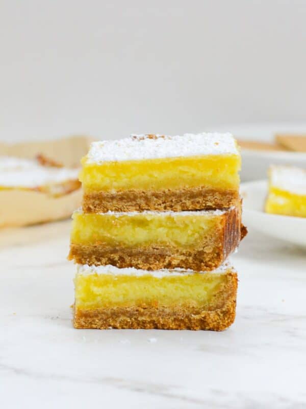 Three sliced and stacked lemon bars with powdered sugar. In the background sit a plate of lemon bars and parchment with sliced bars.