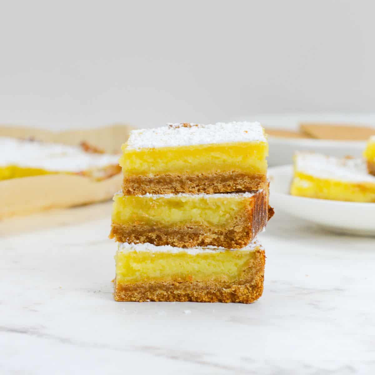 Three sliced and stacked lemon bars with powdered sugar. In the background sit a plate of lemon bars and parchment with sliced bars.
