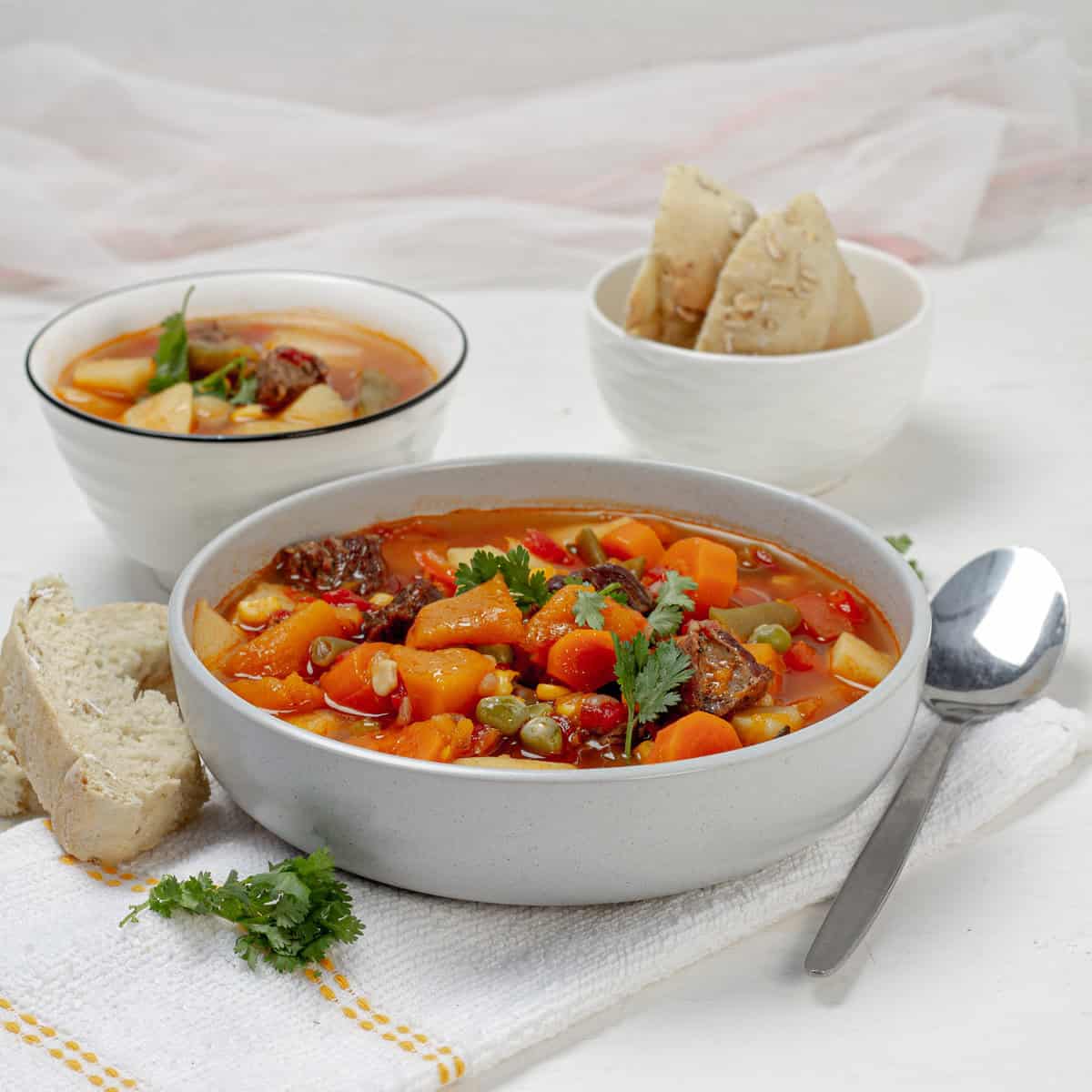 grey bowl of vegetable beef soup with chunks of beef, carrot, potato, pumpkin and peas in tomato broth, a spoon and bread beside it