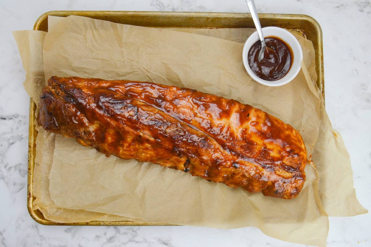An overhead of the finished rack of ribs on a brown parchment-lined baking sheet. A small bowl of sauce and a spoon sit to the side 